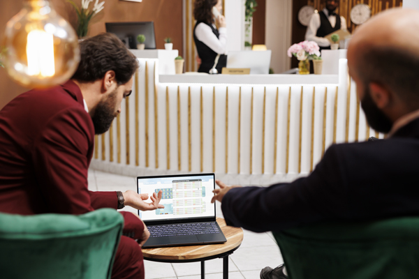 Deux professionnels de l'hôtellerie analysent un tableau de bord de gestion énergétique sur un ordinateur portable dans le hall d'entrée d'un hôtel moderne.