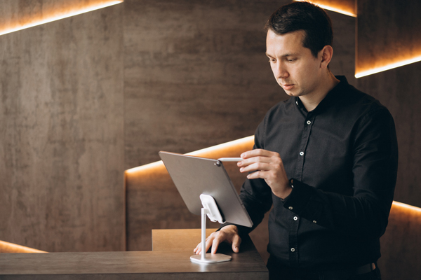 Un exploitant d'hôtel ou gestionnaire utilise un stylet sur une tablette tactile fixée à la réception pour consulter les indicateurs de performance énergétique de son établissement.